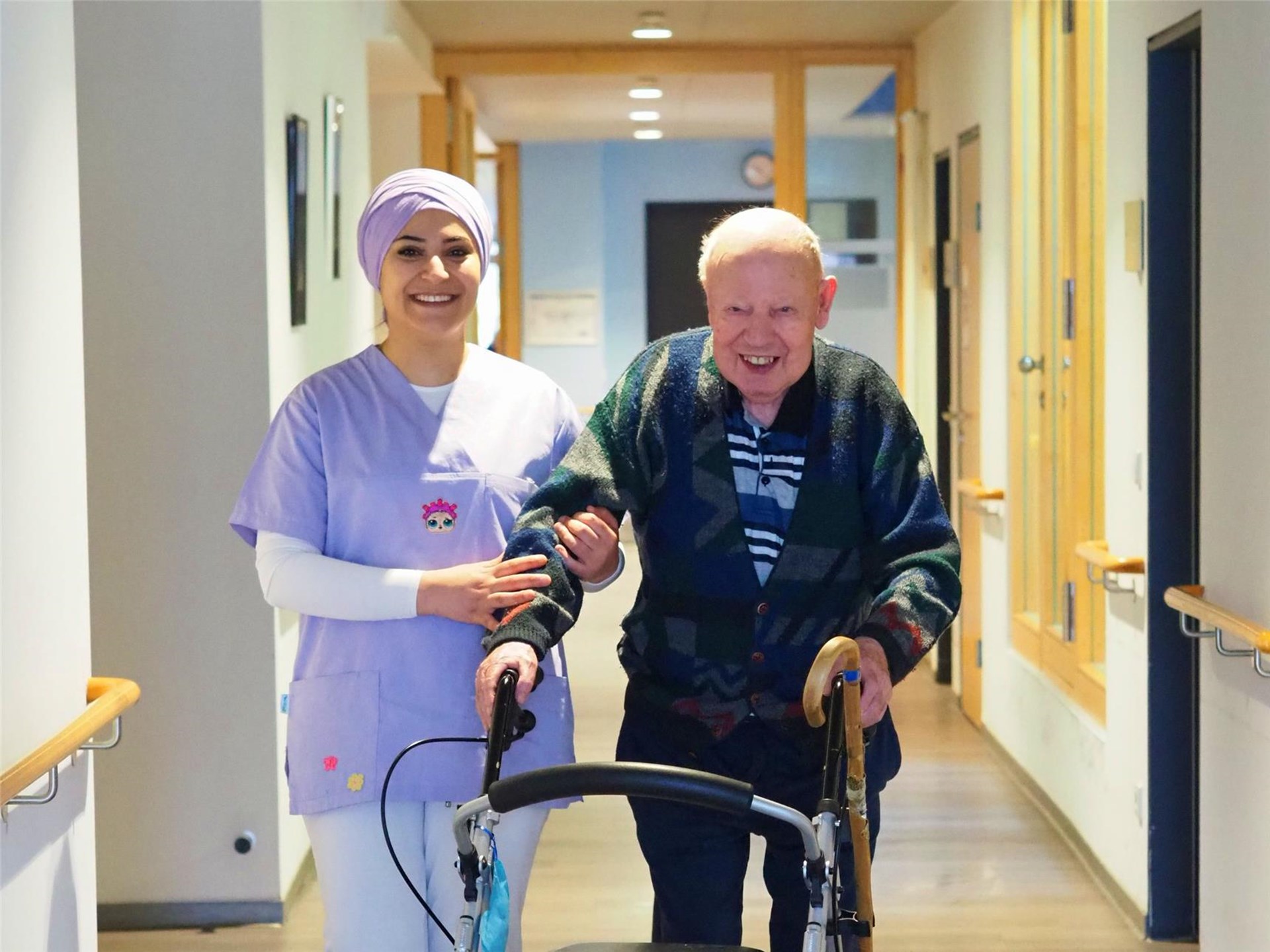 Eine Pflegerin stützt einen Senior, der am Rollator läuft. Sie laufen durch einen Flur. Beide lächeln.