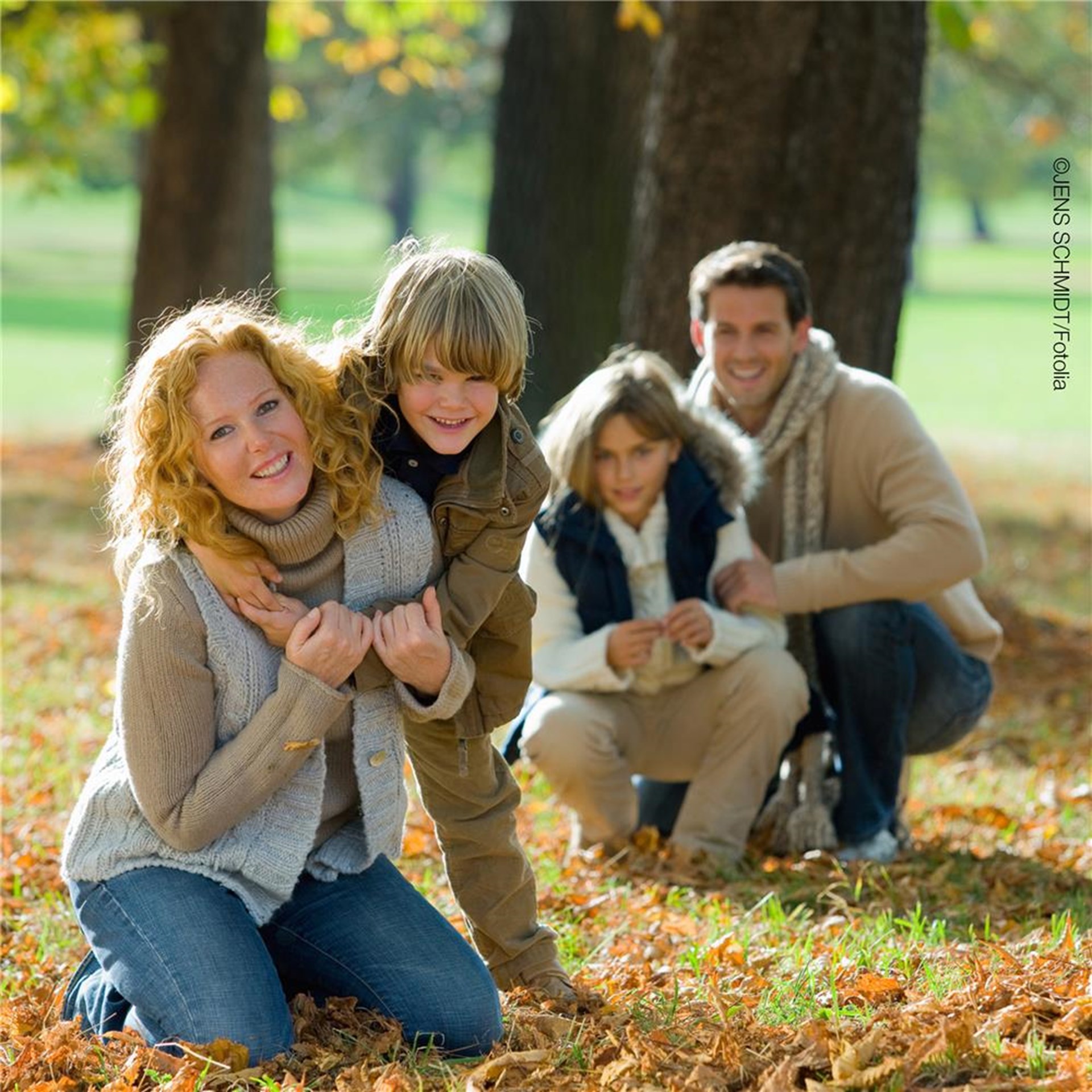 Im Vordergrund eine auf dem Rasen sitzende Frau und ein Junge der sie von hinten umarmt. Im Hintergrund ein Mann und ein weiteres Kind. Es liegt viel Laub auf dem Boden und alle sind warm angezogen.