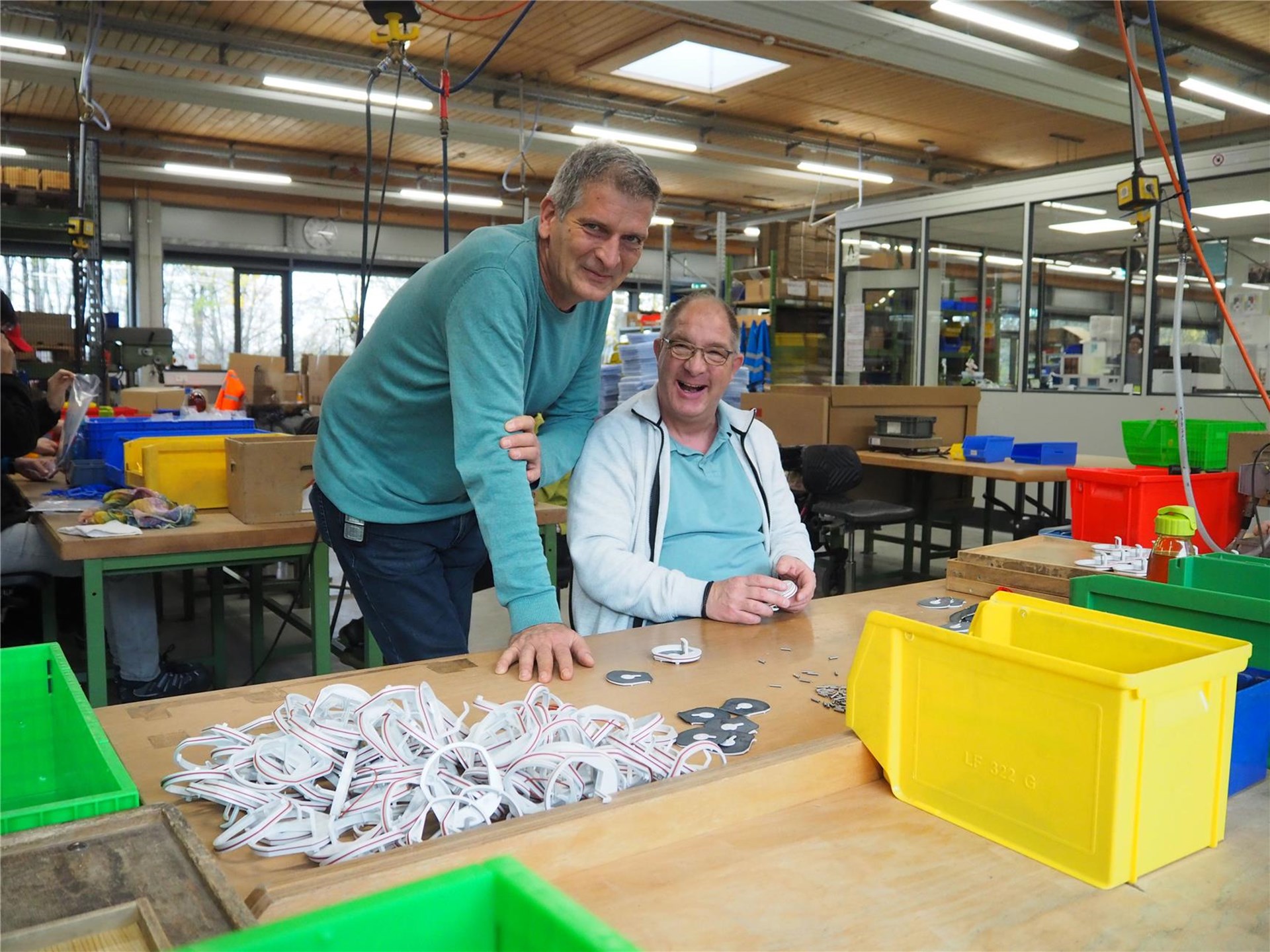 Ein Mitarbeiter neben einem Beschäftigten in der Werkstatt. Der Beschäftigte sitzt an seinem Arbeitsplatz. Vor ihm verschiedene Boxen mit Materialien.